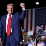 Executive Order Targets SCAM Networks — Finally! A man in a suit raises his fist at a political rally with an American flag in the background