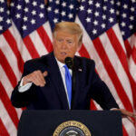 Trump Warns “20X Harder” Strikes A man in a suit speaking at a podium with American flags in the background