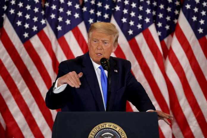 A man in a suit speaking at a podium with American flags in the background