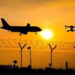 An airplane and a drone flying against a sunset backdrop with a barbed wire fence