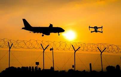 shutterstock_1263858730.jpg An airplane and a drone flying against a sunset backdrop with a barbed wire fence