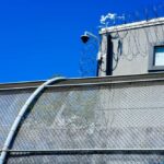Massive Trial: Protesters or Terrorists at ICE? Security camera mounted above a prison fence with barbed wire against a blue sky