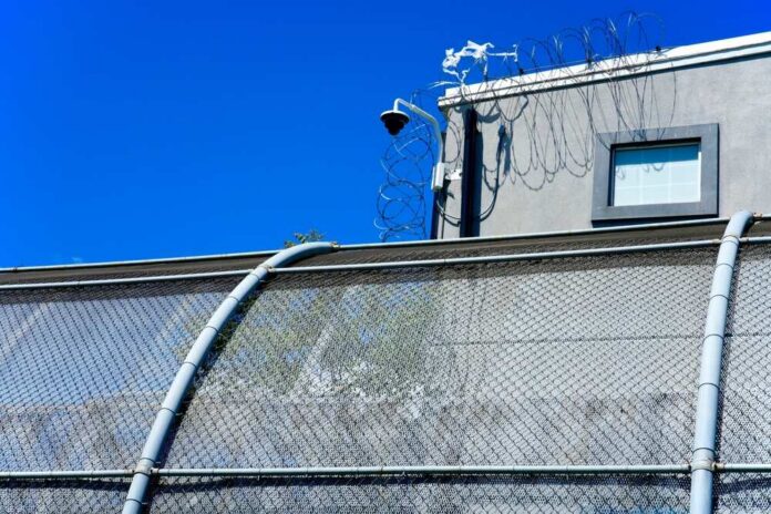 shutterstock_2625726951.jpg Security camera mounted above a prison fence with barbed wire against a blue sky