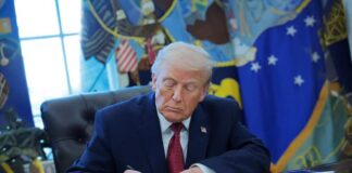 A person in a suit signing documents at a desk in the Oval Office