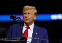 A man in a blue suit and red tie speaking into a microphone on stage