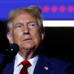 A man in a suit speaking into a microphone with a serious expression