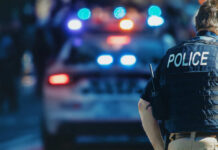 Police officer in a tactical vest standing in front of a police car with flashing lights