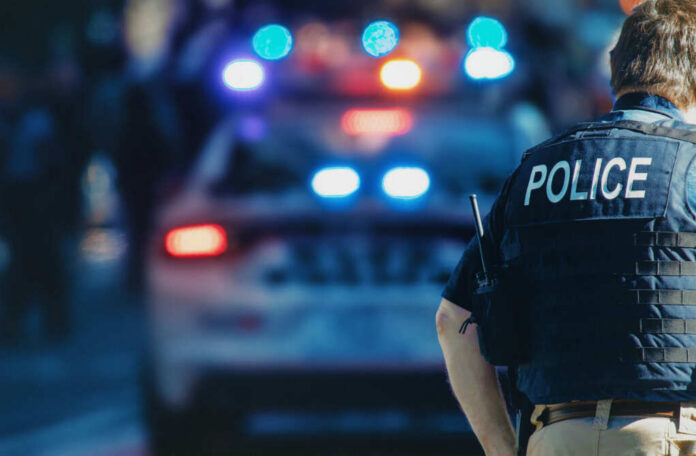 shutterstock_1543591088.jpg Police officer in a tactical vest standing in front of a police car with flashing lights