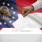 Democrat Dreams DIE in Georgia Bloodbath A hand placing a ballot into a ballot box with the Georgia state flag in the background