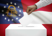 Democrat Dreams DIE in Georgia Bloodbath A hand placing a ballot into a ballot box with the Georgia state flag in the background