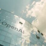 A modern glass building with the OpenAI logo against a cloudy sky
