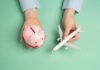 Hands holding a piggy bank and a toy airplane against a mint green background