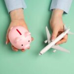 Hands holding a piggy bank and a toy airplane against a mint green background