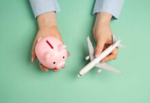 Hands holding a piggy bank and a toy airplane against a mint green background