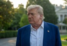 A political figure with a serious expression standing outdoors near the White House