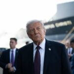 A political figure in a suit looking thoughtfully at an outdoor event with a ship in the background