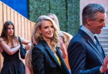 Two individuals in formal attire at a red carpet event, with others in the background