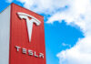 Tesla logo on a red corporate sign against a blue sky with clouds