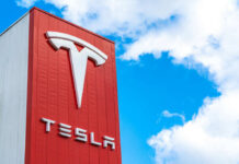 Tesla logo on a red corporate sign against a blue sky with clouds