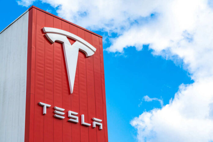 Tesla logo on a red corporate sign against a blue sky with clouds