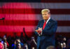 A man in a suit stands in front of an American flag, applauding at a rally