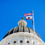 View of a state capitol dome with American and California flags against a blue sky