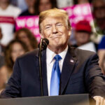 Mysterious C-SPAN Caller Sparks Trump Revolt Buzz A speaker smiling at a political rally with a microphone in front of him and an engaged audience behind.