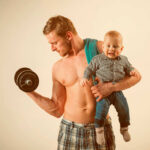 Internet EXPLODES Over Parenting Debate A muscular man lifting a dumbbell while holding a smiling baby