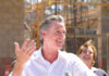 A man speaking at a construction site with a microphone, smiling, while a woman in the background laughs.