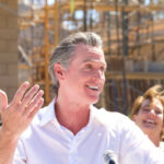Explosive Allegations: Newsom’s Cozy Diaper Deal A man speaking at a construction site with a microphone, smiling, while a woman in the background laughs.