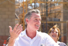 Explosive Allegations: Newsom’s Cozy Diaper Deal A man speaking at a construction site with a microphone, smiling, while a woman in the background laughs.