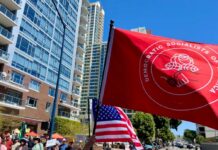 Protesters march in a city street holding a large red flag and U.S. flags