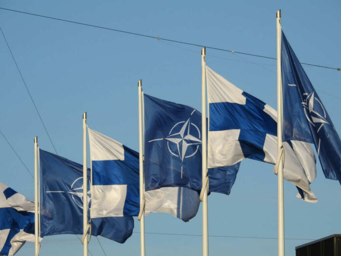 Flags of Finland and NATO waving in the wind against a clear blue sky