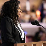 A woman in formal attire speaking at a podium with microphones