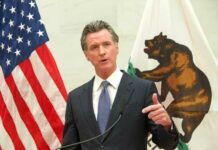 Questions Grow Around Newsom Probe A government official delivering a speech with flags in the background