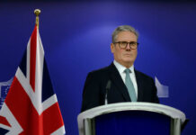 A political leader standing at a podium with a British flag in the background