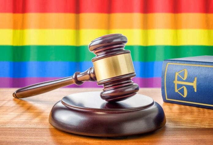 A,Gavel,And,A,Law,Book,-,Rainbow,Flag A wooden gavel on a table with a rainbow flag in the background