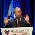 A speaker gesturing during a political conference with flags in the background