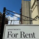 Housing Costs Meet Immigration Reality Sign indicating apartments available for rent against a blue sky