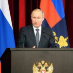 Trash Bin Bomb Kills Former Commander man in suit speaking at a podium with flags behind him