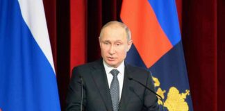 man in suit speaking at a podium with flags behind him