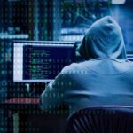 Person in hoodie working at computer with code on screens