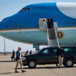 Security detail near blue and white aircraft with staircase and SUV