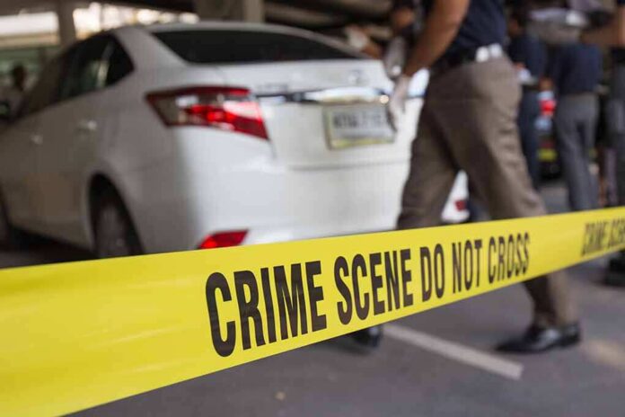 shutterstock_351048443 Yellow crime scene tape in front of a white car with investigators nearby