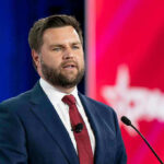 Vance Drops Fraud Hammer – Who Gets Cut? Man in suit speaking at a podium with microphone