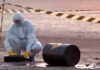 Hazmat worker kneeling near a fallen barrel at a cordoned-off site