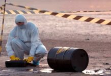Hazmat worker kneeling near a fallen barrel at a cordoned-off site