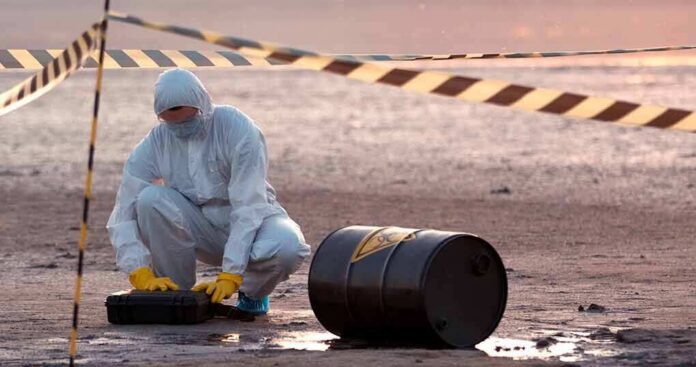 Hazmat worker kneeling near a fallen barrel at a cordoned-off site
