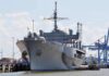 Large gray naval ship docked at an industrial port